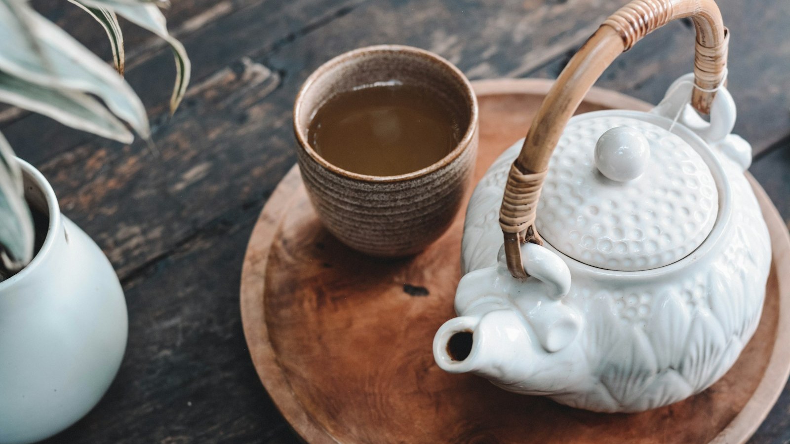 A stoneware cup of pale herbal tea beside a white ceramic teapot on a wooden tray