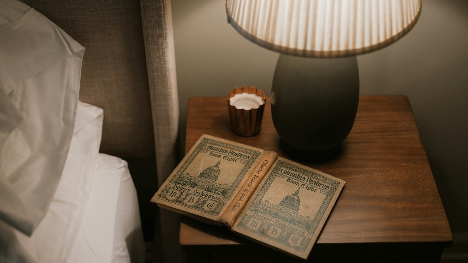 An old cloth-bound book lying open on a wooden bedside table beside a lit lamp